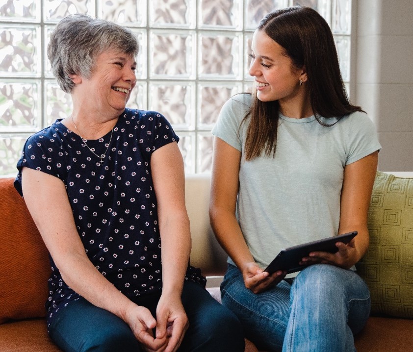 A young woman is smiling and holding a tablet using the LifeBio Memory app while her grandmother smiles and reminisces her life memories